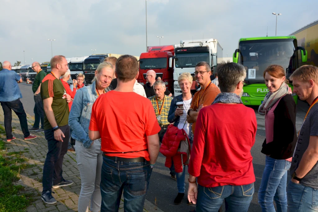 Gruppe von Menschen im Gespräch auf einem Parkplatz vor LKWs und einem Bus bei sonnigem Wetter.
