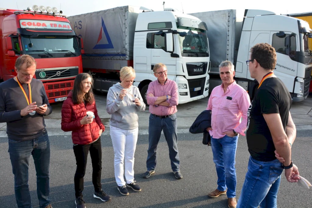 Gruppe von sechs Personen, die sich vor Lastwagen unterhalten, während einige Getränke halten. Im Hintergrund sind verschiedene Lkw-Modelle sichtbar, darunter ein roter Volvo und ein weißer MAN.