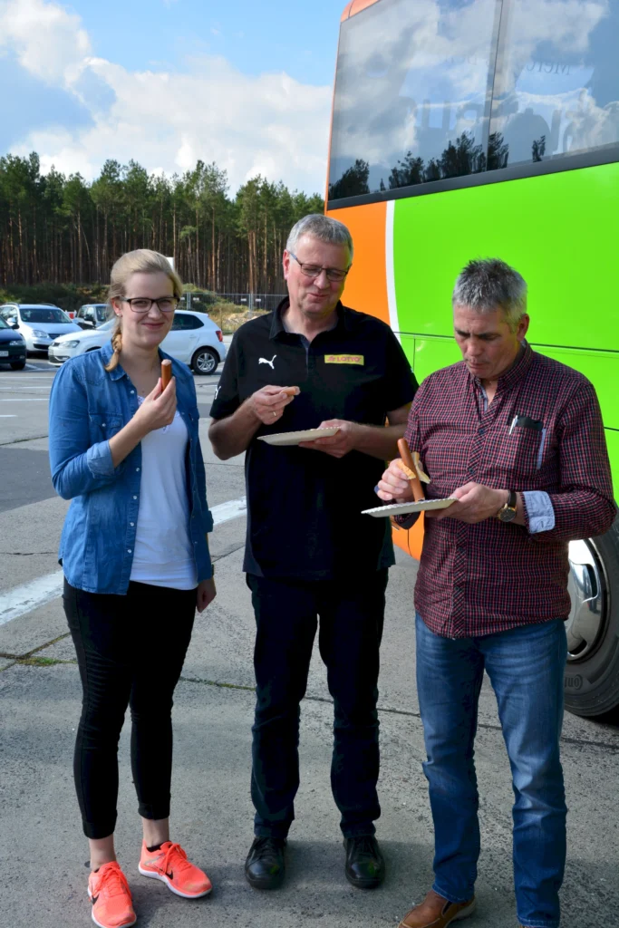 Drei Personen essen Würstchen vor einem grünen Bus auf einem Parkplatz, umgeben von Autos und Bäumen.
