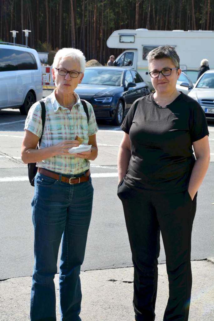 Zwei Personen stehen auf einem Parkplatz im Freien vor Autos. Eine Person hält einen Snack in der Hand.