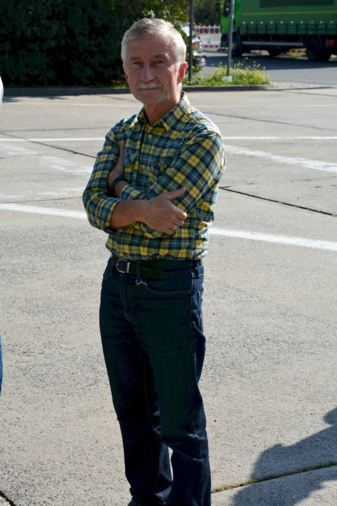 Älterer Mann mit verschränkten Armen, kariertes Hemd, steht draußen auf Asphalt, im Hintergrund Bäume und LKW.