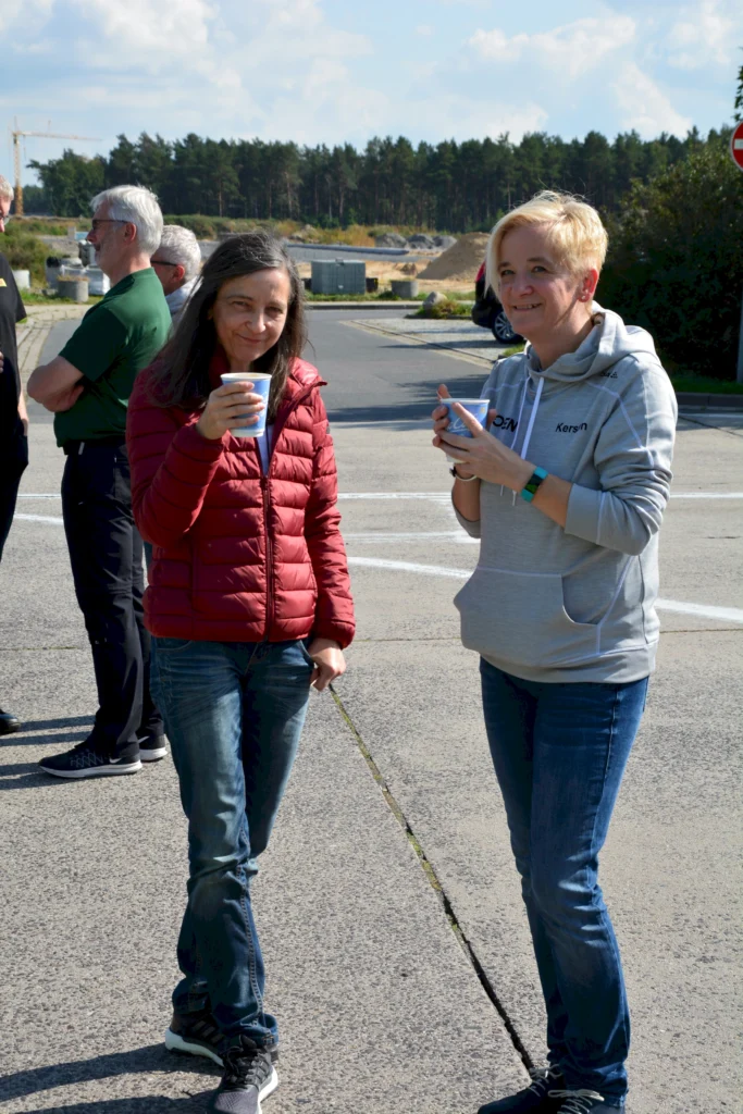 Zwei Frauen trinken draußen Kaffee, umgeben von Menschen. Entspannte Atmosphäre bei Sonnenschein.