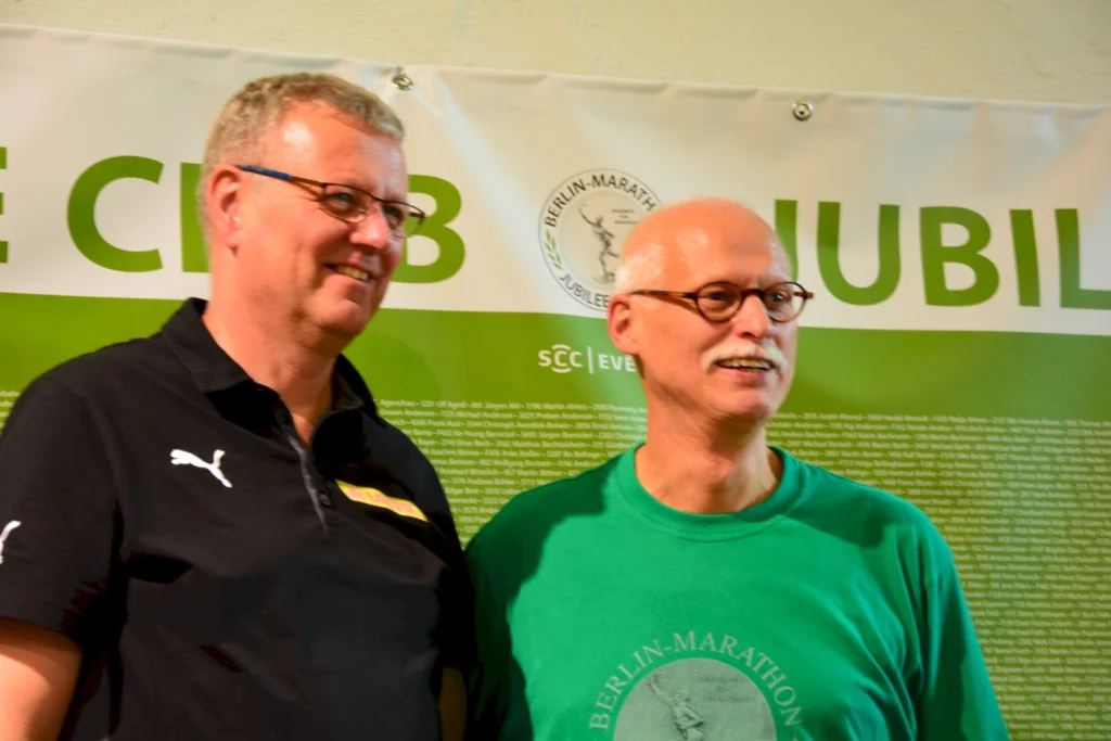 Zwei Männer bei einer Veranstaltung des Berlin-Marathons vor einem Jubiläumsbanner.