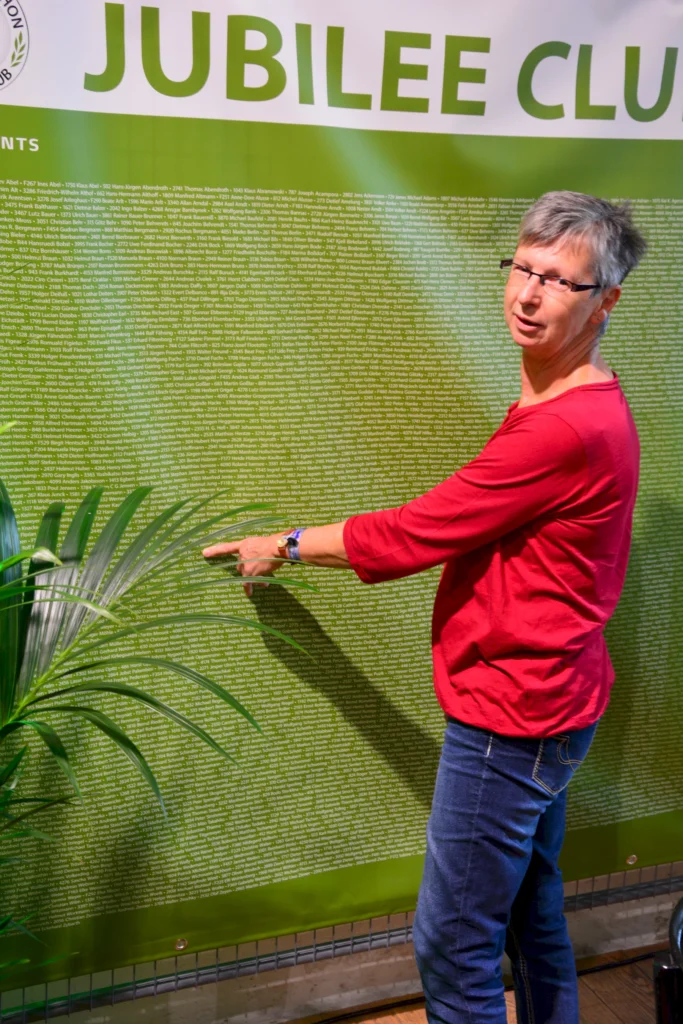 Person zeigt auf eine grüne Jubiläumswand mit vielen Namen. Text: JUBILEE CLUB. Links eine Pflanze.