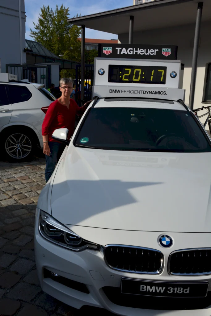 Person mit rotem Oberteil neben weißem BMW 318d mit digitaler Uhranzeige von 20:17 und TAG Heuer Logo.