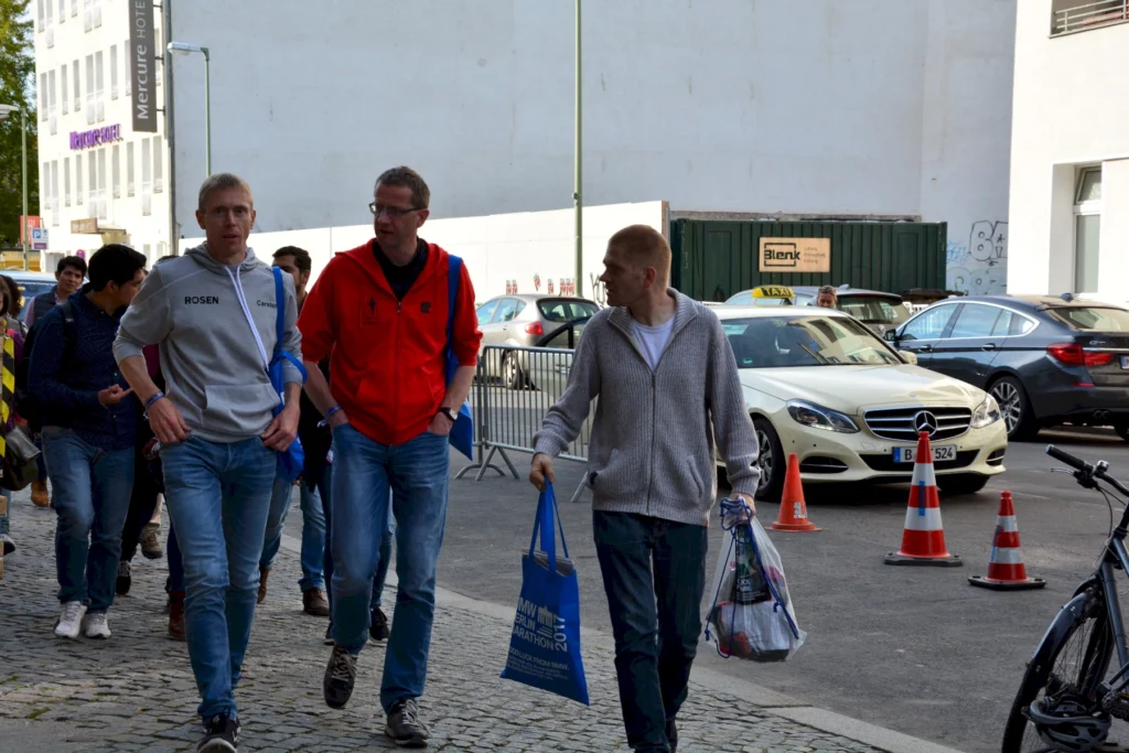 Gruppe von Menschen geht auf Gehweg neben Autos in der Stadt, zwei von ihnen tragen Taschen, im Hintergrund ein Taxi.