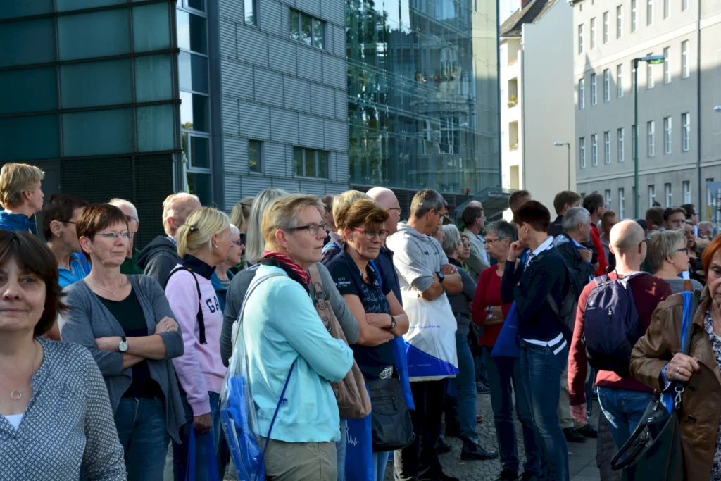 Gruppe von Menschen versammelt sich vor modernen Gebäuden bei Tageslicht, alle tragen Freizeitkleidung und beobachten etwas.