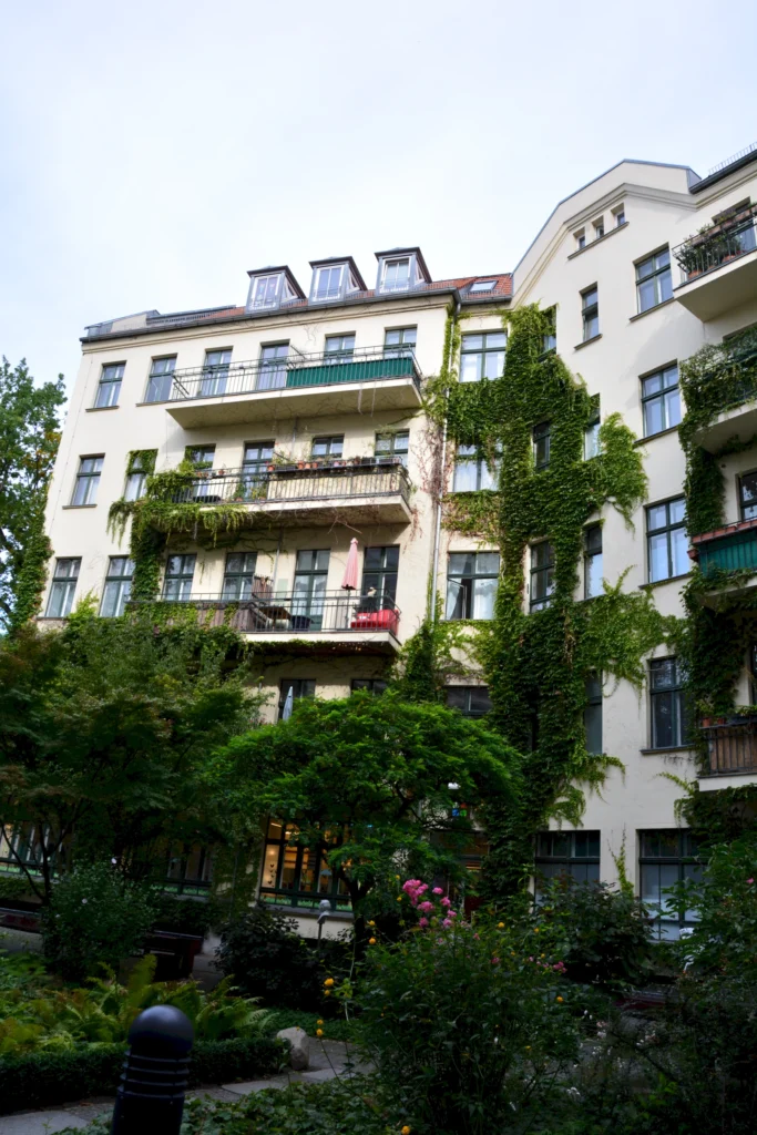 Mehrstöckiges Wohngebäude mit Balkonen, teils von Efeu bewachsen, umgeben von grüner Gartenanlage.