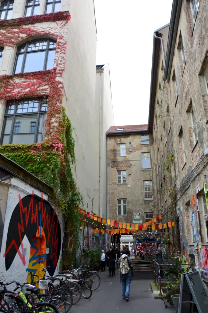 Belebte Berliner Gasse mit Graffiti und Fahrrädern, geschmückt mit bunten Lichtern und Menschen.