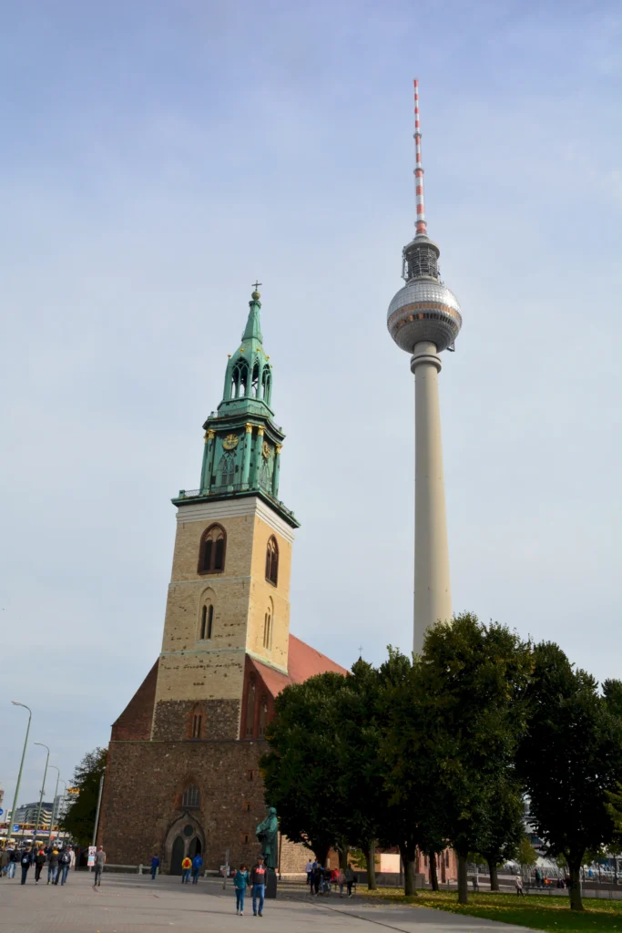 Blick auf die Marienkirche und den Fernsehturm in Berlin, umgeben von Menschen und Bäumen, mit einem bewölkten Himmel im Hintergrund.