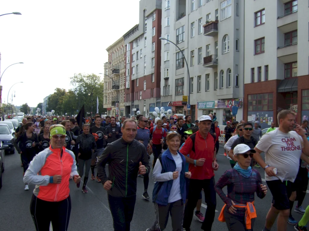 Gruppe von Läufern bei einem Straßenlauf in einer Stadt, umgeben von hohen Gebäuden und Zuschauerautos.