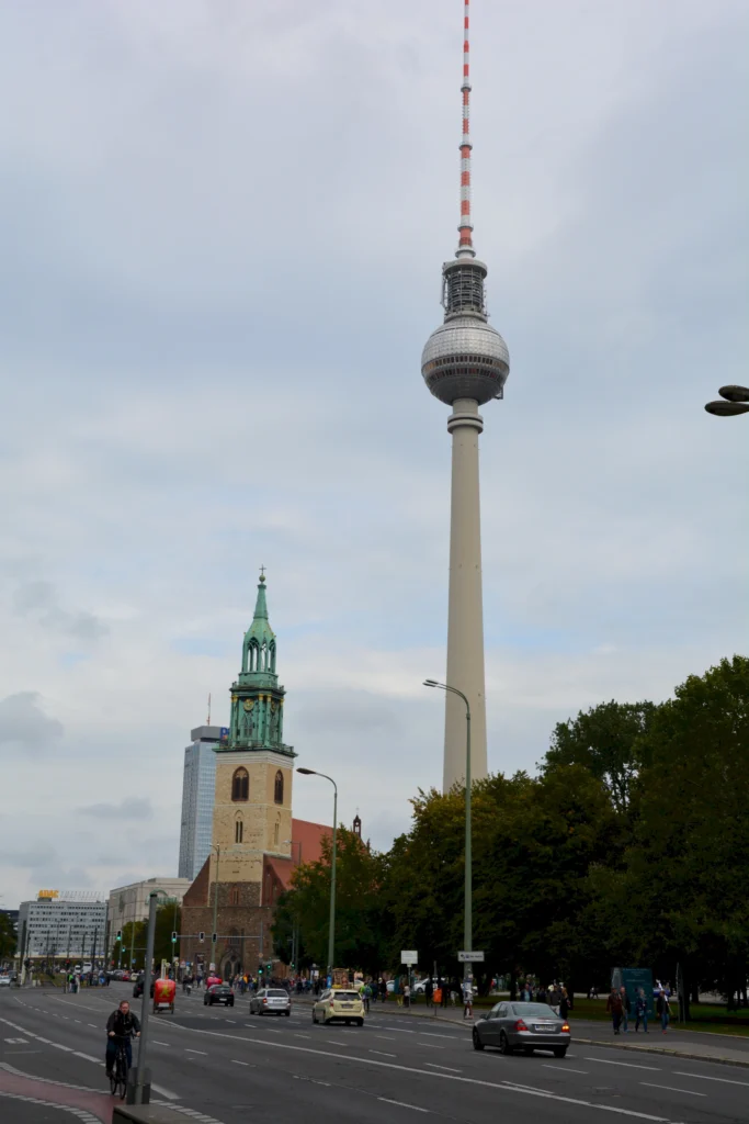 Berliner Fernsehturm und Marienkirche vor bewölktem Himmel, Autos und Fahrräder auf der Straße im Vordergrund.