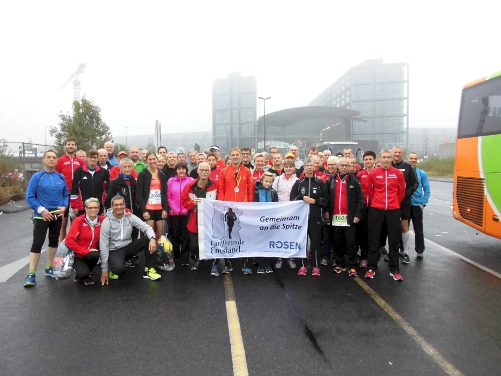 Gruppe von Läufern in Sportkleidung steht vor modernem Gebäude und Bus, hält Banner mit Vereinsname und Slogan.