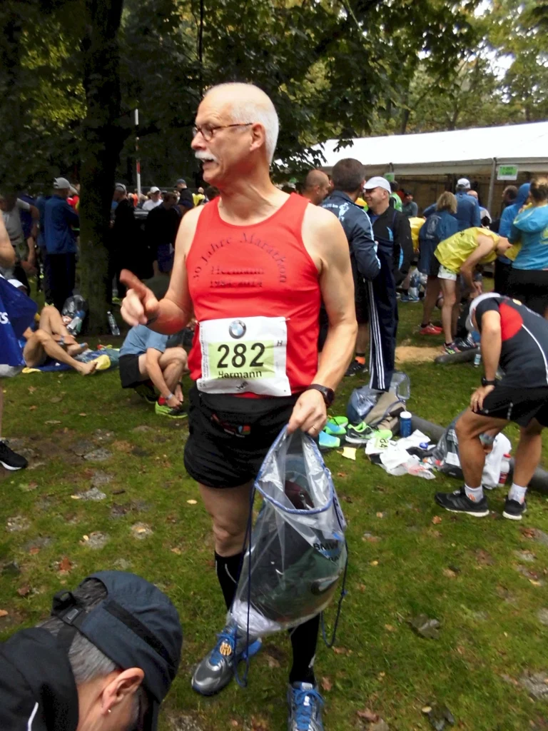 Mann im roten Laufshirt bereitet sich auf Marathon im Park vor; hält Tasche, umgeben von anderen Läufern.