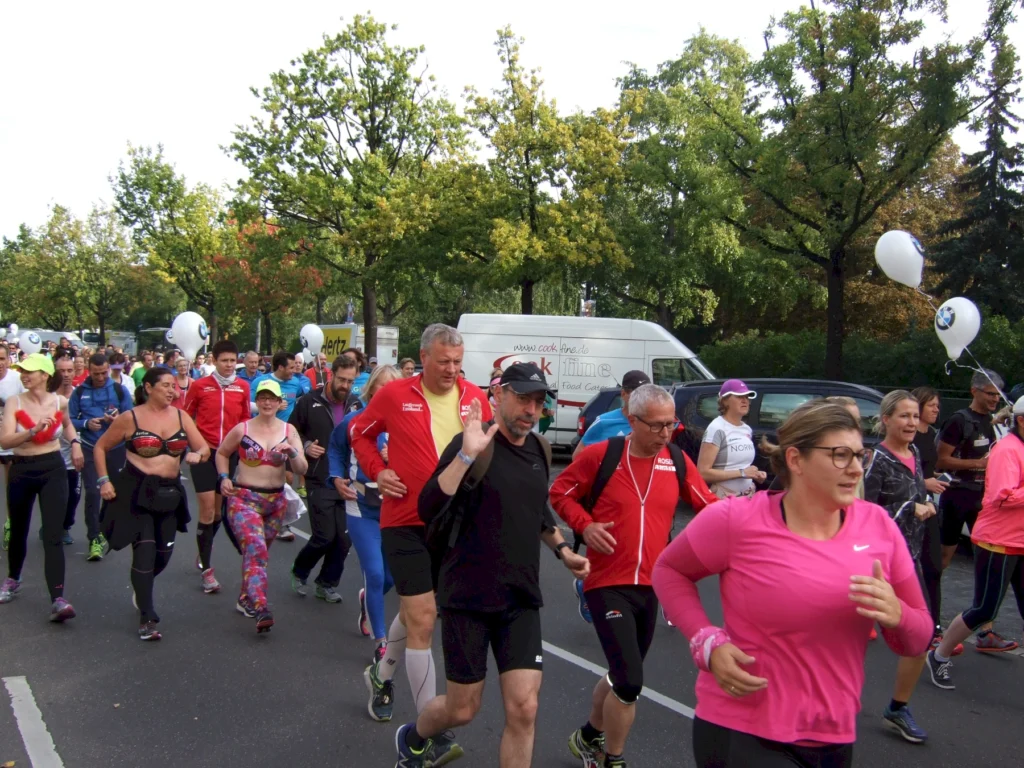Läufergruppe bei Stadtmarathon im Park, bunte Sportkleidung, Bäume im Hintergrund, sonniger Tag, gemeinsamer Laufspaß.