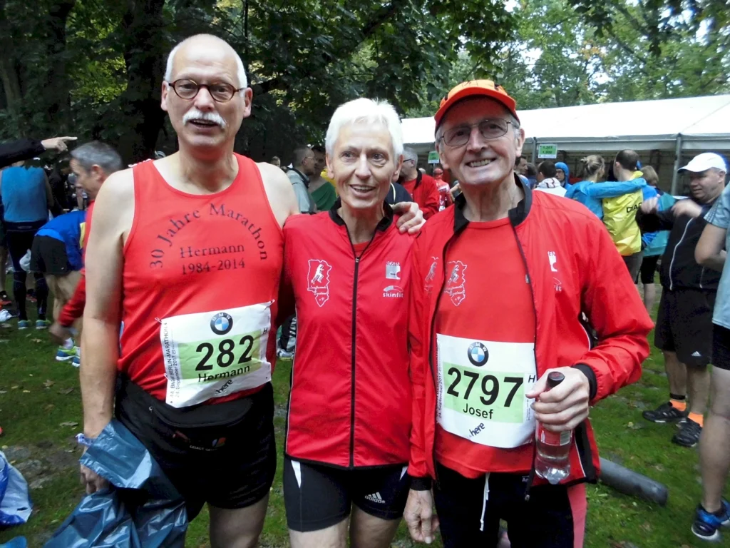 Drei Läufer stehen vor einem Marathon-Event, gekleidet in rote Trikots. Der Mann links trägt ein Shirt mit der Aufschrift "30 Jahre Marathon", während die Frau in der Mitte und der Mann rechts ebenfalls sportliche Kleidung mit Nummern haben. Im Hintergrund sind weitere Teilnehmer zu sehen, die sich auf das Rennen vorbereiten.