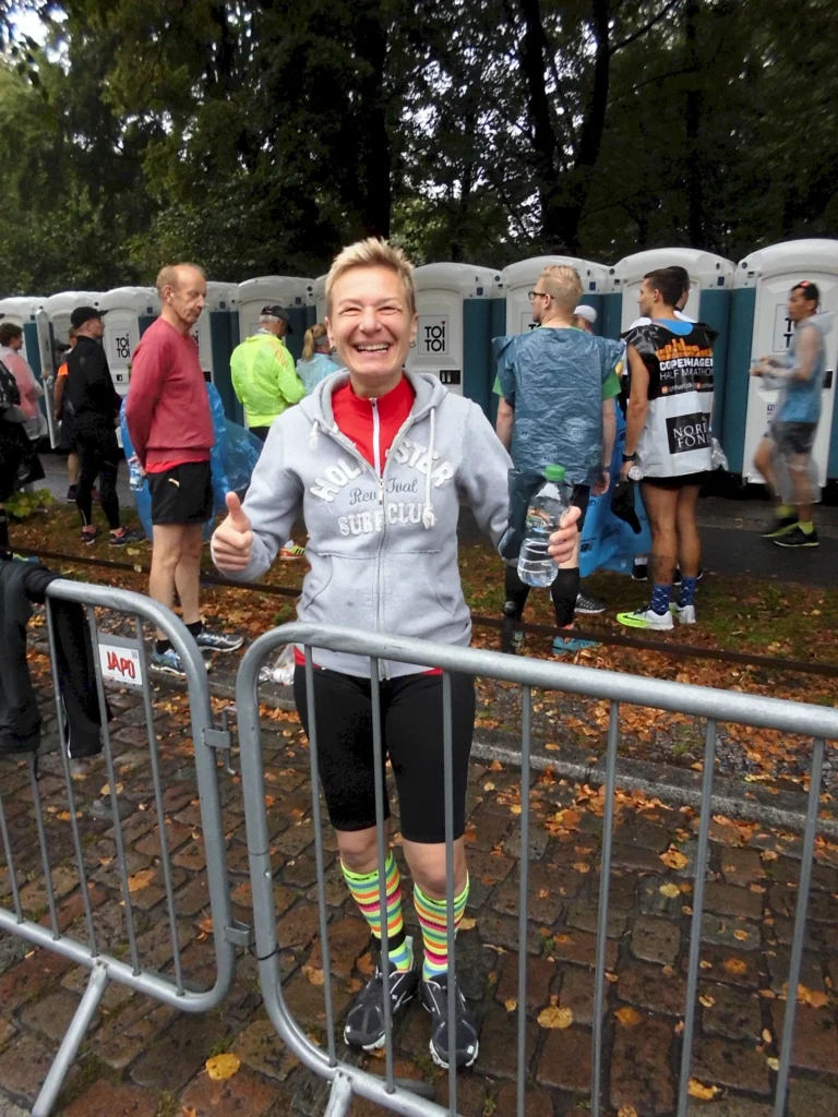Lächelnde Teilnehmerin mit bunten Socken beim Marathon, zeigt Daumen hoch vor mobilen Toilettenanlagen im Freien.