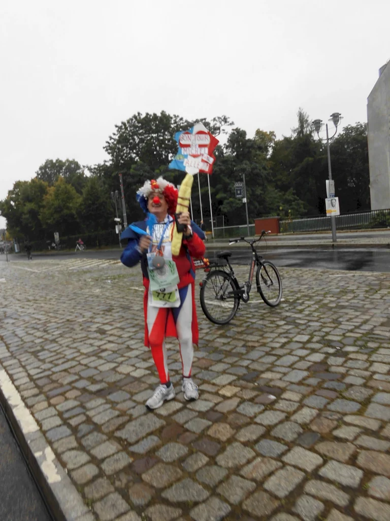 Person in bunter Verkleidung mit Schildern und Fahrrad auf Kopfsteinpflasterstraße bei bewölktem Himmel.