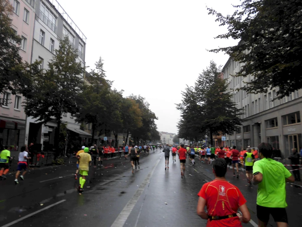 Teilnehmer eines Straßenrennens laufen an einer regnerischen Straße entlang, umgeben von Bäumen und Gebäuden. Viele Läufer tragen bunte Sportkleidung, darunter leuchtendes Gelb und Rot.