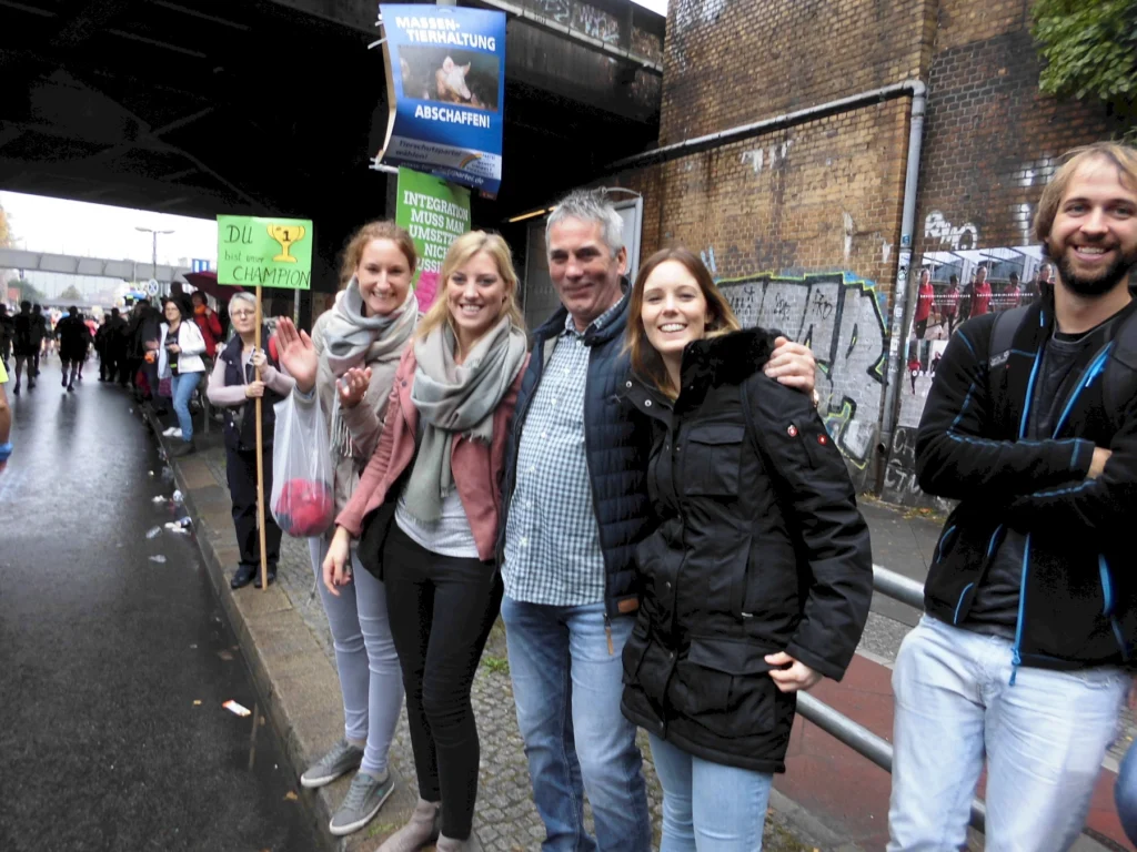 Vier Personen stehen unter einer Brücke und lächeln während einer Demonstration. Im Hintergrund sind Plakate mit Tierschutzforderungen zu sehen. Die Atmosphäre ist lebhaft und engagiert, mit weiteren Demonstrationsteilnehmern im Hintergrund.