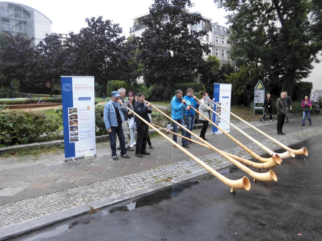 Gruppe von Musikern, die Alphörner spielen, während sie an einer Veranstaltung der Leo-Kestenberg-Musikschule teilnehmen. Im Hintergrund sind Informationsbanner und eine parkähnliche Umgebung sichtbar.