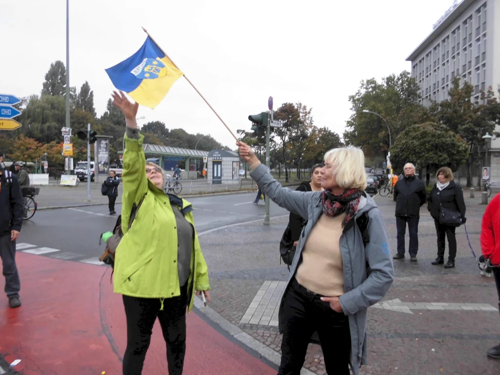 Personen auf Straße schwenken blau-gelbe Flagge in einer Stadtumgebung, Bäume und Gebäude im Hintergrund.