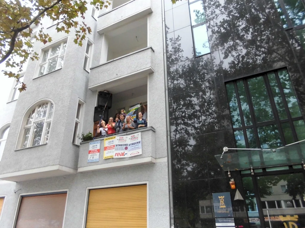 Menschen stehen auf einem Balkon eines grauen Mehrfamilienhauses und winken, während im Hintergrund ein modernes Gebäude mit reflektierenden Fenstern zu sehen ist. Auf dem Balkon hängen mehrere Plakate mit Informationen zu einem Event oder einer Veranstaltung.