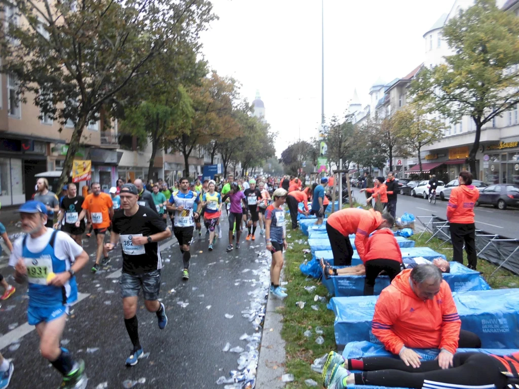 Läufer beim Berlin Marathon in einer regnerischen Umgebung, während Helfer an der Strecke Unterstützung bieten. Die Straße ist mit Wasserflaschen und Abfall bedeckt, und die Teilnehmer tragen bunte Sportkleidung.