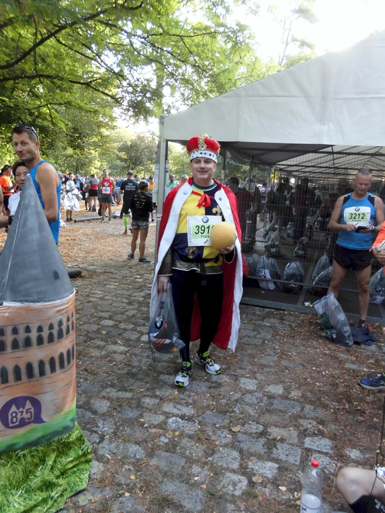 Teilnehmer eines Marathons verkleidet als König mit Krone und Umhang, steht vor einem Zelt unter grünen Bäumen.