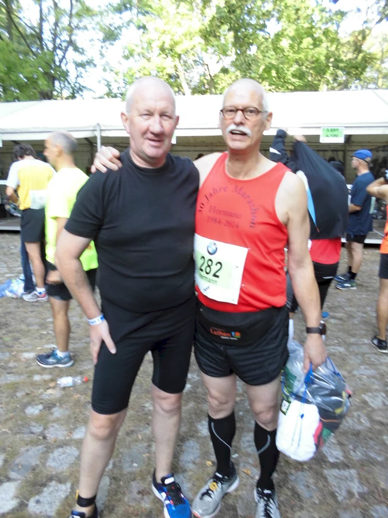 Zwei Männer, die in Sportkleidung bei einem Marathon-Event im Freien stehen, umgeben von anderen Teilnehmern.