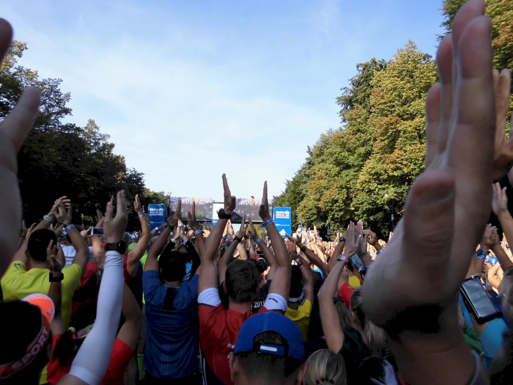 Läufer jubeln beim Marathonstart im Park bei sonnigem Wetter, umgeben von Bäumen und blauen Schildern.