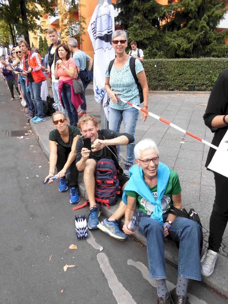 Menschenmenge jubelt bei Sportveranstaltung auf Gehweg, einige halten Kameras und Schirme, fröhliche Stimmung.