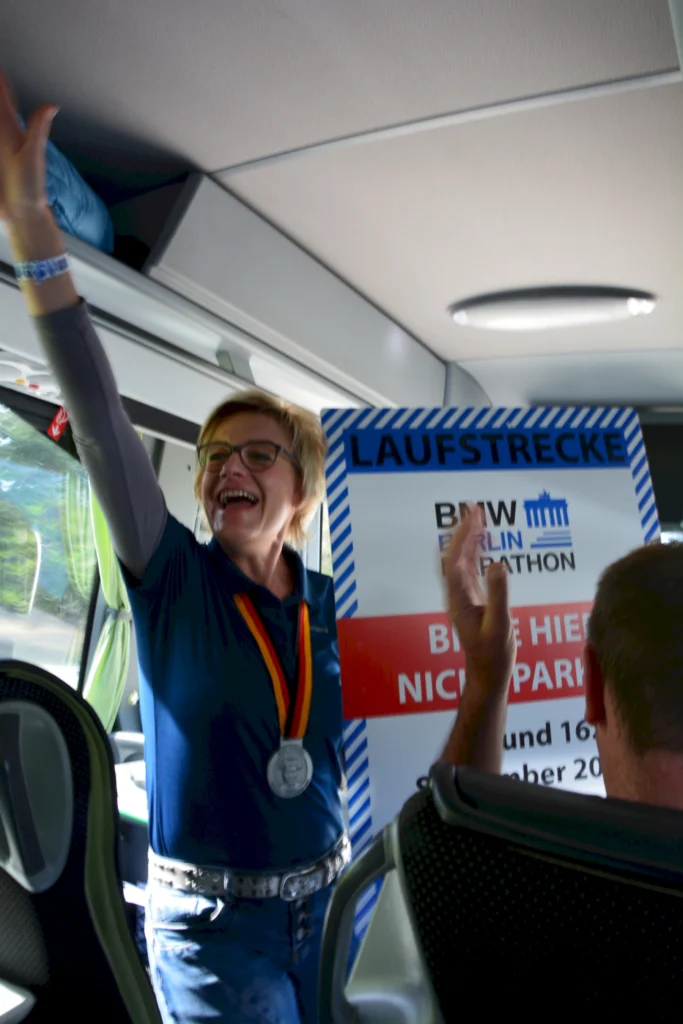 Frau im Bus mit Medaille, hält Marathon-Plakat, lächelnd und feiernd, in einer fröhlichen Atmosphäre.