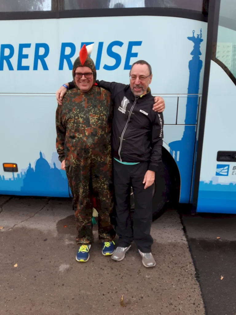 Zwei Männer in Kostüm und Freizeitkleidung lächeln vor einem Reisebus und umarmen sich.