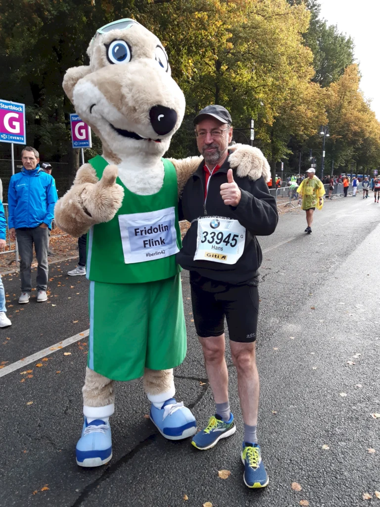 Läufer posiert mit einem Maskottchen bei einem Straßenlauf im Herbst, gibt Daumen hoch, umgeben von Bäumen und Zuschauern.
