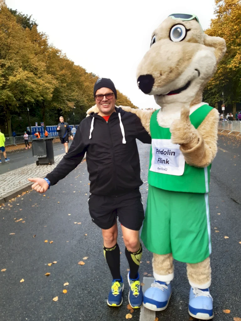 Läufer posiert mit einem Maskottchen in einem Park während eines Lauf-Events, umgeben von herbstlichen Bäumen und anderen Teilnehmern im Hintergrund.
