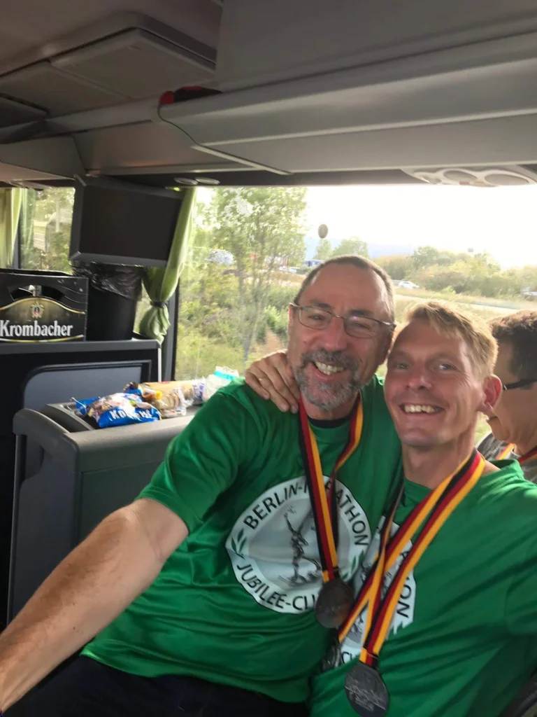 Zwei Männer in grünen Berlin-Marathon-Shirts mit Medaillen lächeln und umarmen sich im Bus.