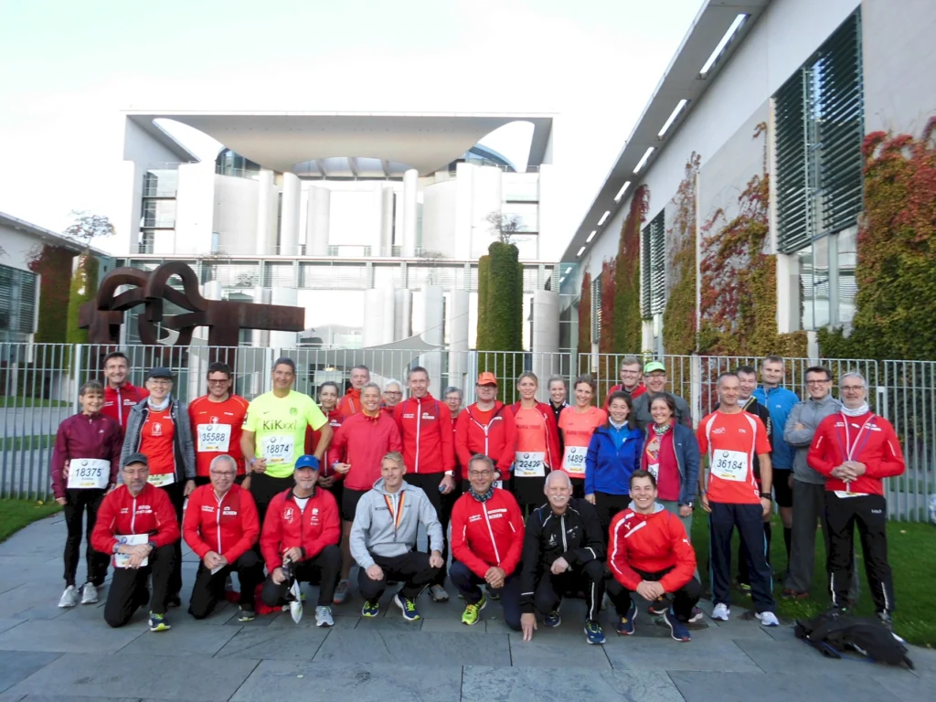 Gruppe von Laufteilnehmern in roten Trikots vor dem Bundeskanzleramt in Berlin, bereit für ein Lauf-Event, mit verschiedenen Laufnummern sichtbar.