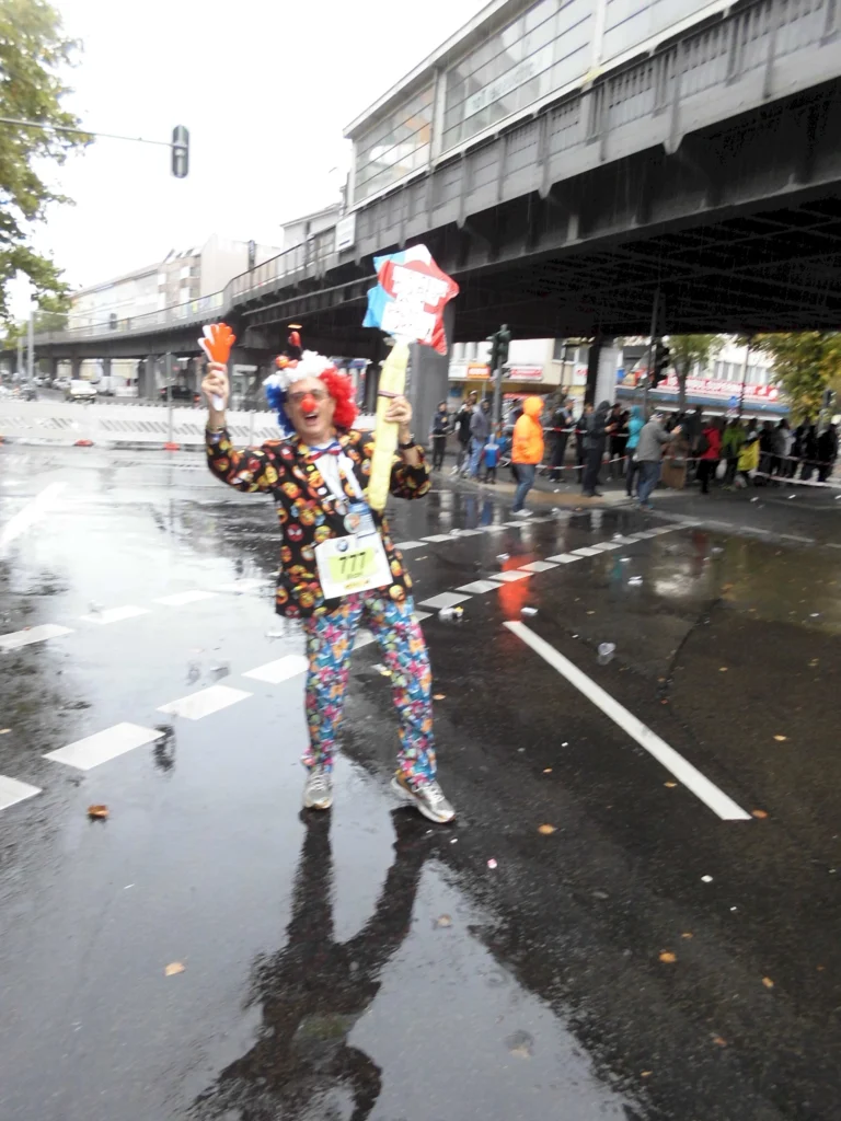 Clown mit buntem Kostüm und Schildern steht auf regennasser Straße während einer Veranstaltung, umgeben von Zuschauern und einem urbanen Hintergrund.