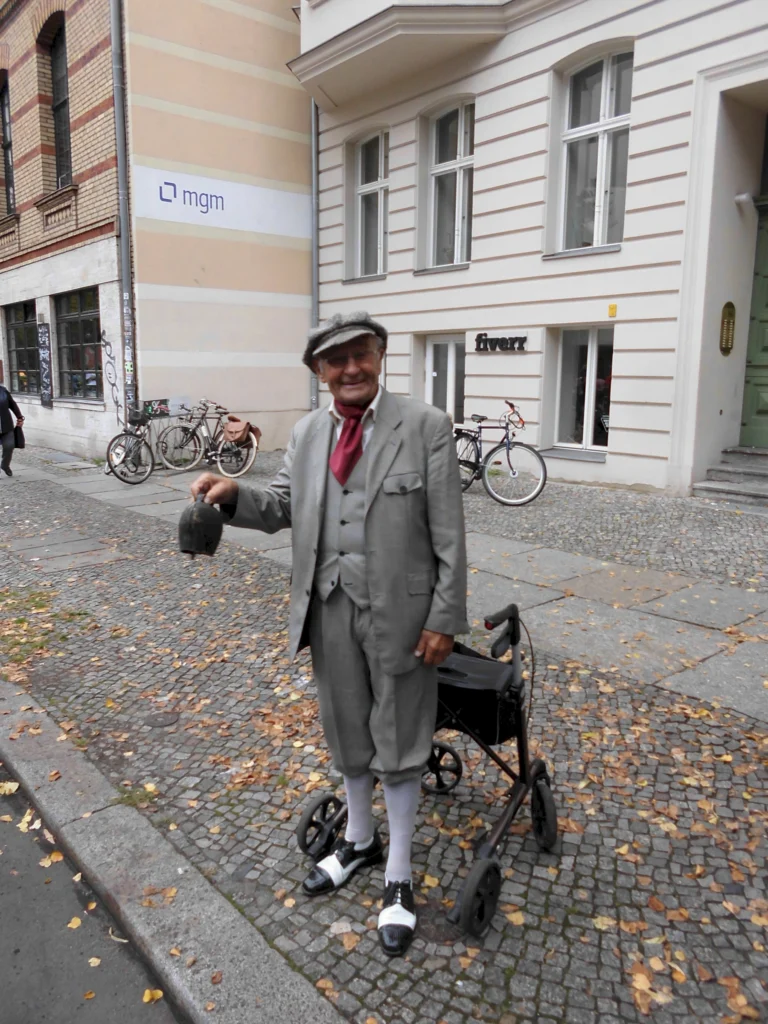 Älterer Herr in grauem Anzug mit Rollator auf Berliner Straße, lächelnd in die Kamera, herbstliche Atmosphäre im Hintergrund.