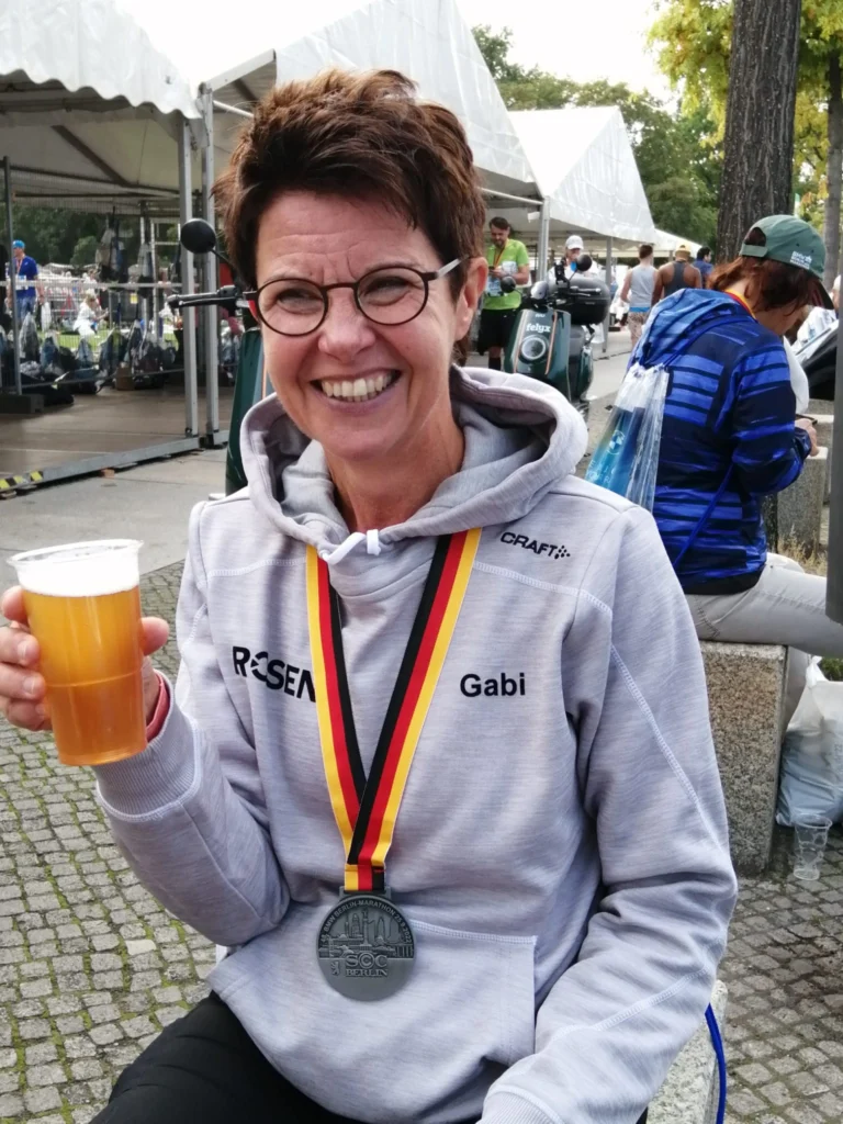 Person in Sportbekleidung mit Marathonmedaille und Getränk in der Hand, lächelnd auf einem Sportevent im Freien.