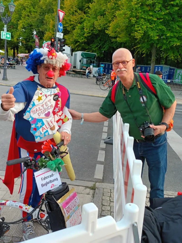 Ein Mann im Clown-Kostüm posiert mit Fahrrad neben Fotografen auf der Straße, umgeben von Bäumen und Schildern.