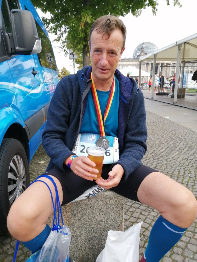 Ein Marathonläufer entspannt sich nach dem Lauf mit einer Medaille und einem Bier in der Hand in einer städtischen Umgebung.