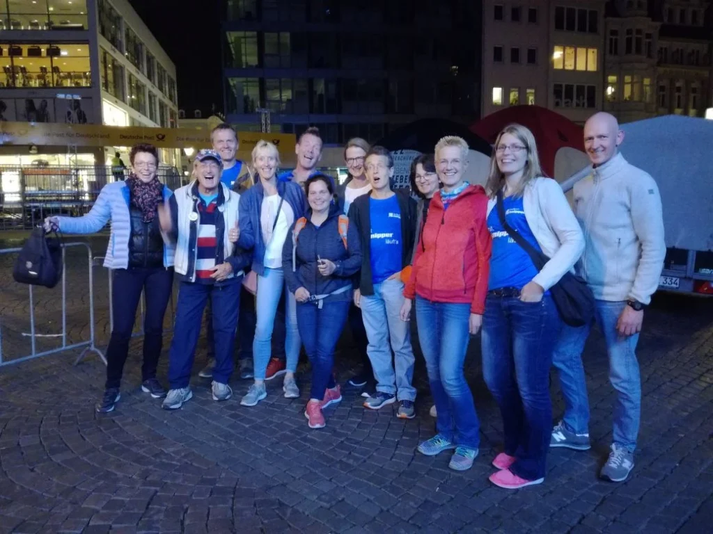 Gruppe von 13 Personen, die in der Nacht fröhlich posieren, vor einem urbanen Hintergrund mit Gebäuden und Lichtern. Einige tragen blaue T-Shirts mit Aufdrucken, während andere lässige Kleidung tragen. Die Stimmung ist ausgelassen und freundlich.