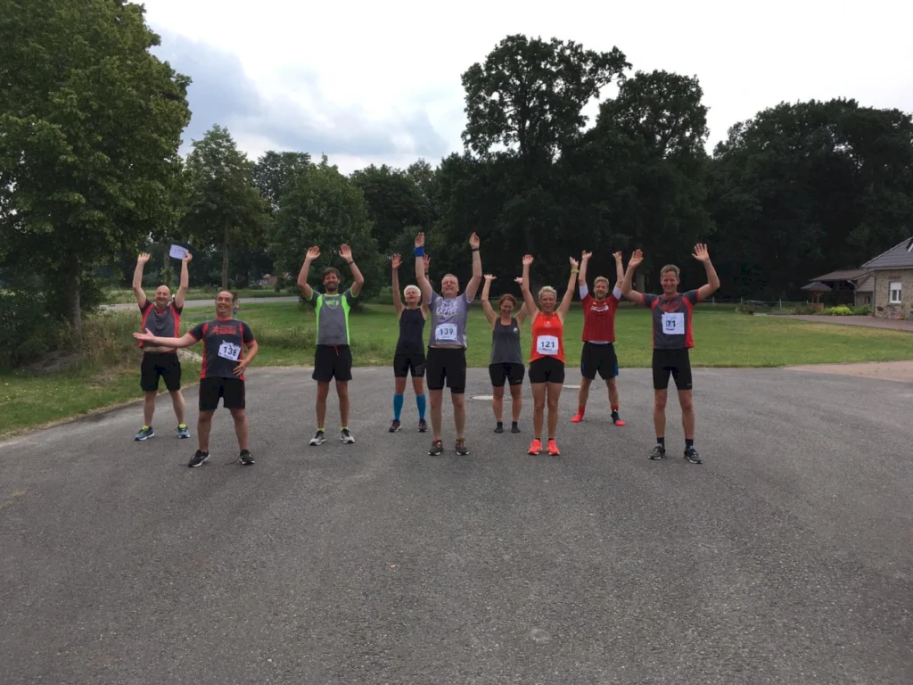 Gruppe von Läufern mit erhobenen Händen, die ihre Nummern tragen, stehen auf einer Straße in einer grünen Umgebung. Ideal für einen Lauf-Event oder ein Sporttraining.