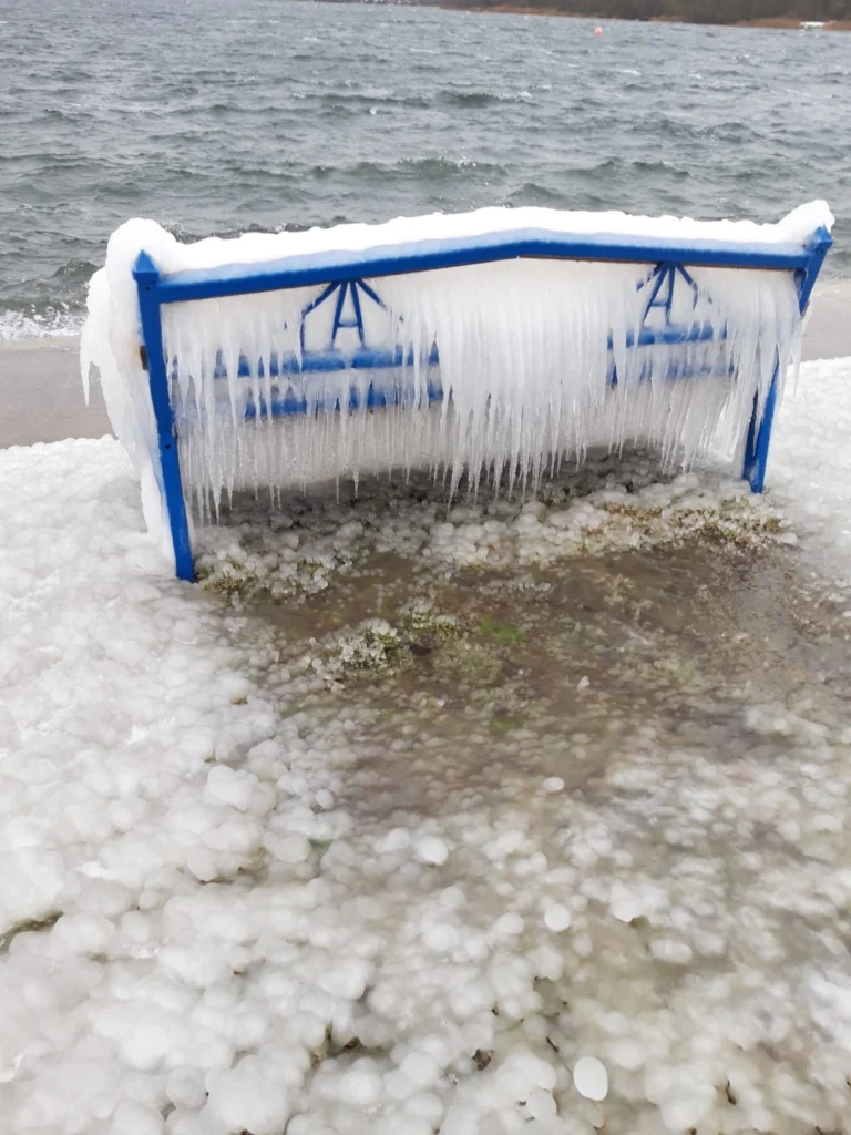 Eingefrorene Bank am Wasser mit Eiszapfen und Schnee, umgeben von gefrorenem Wasser und Eisbläschen.