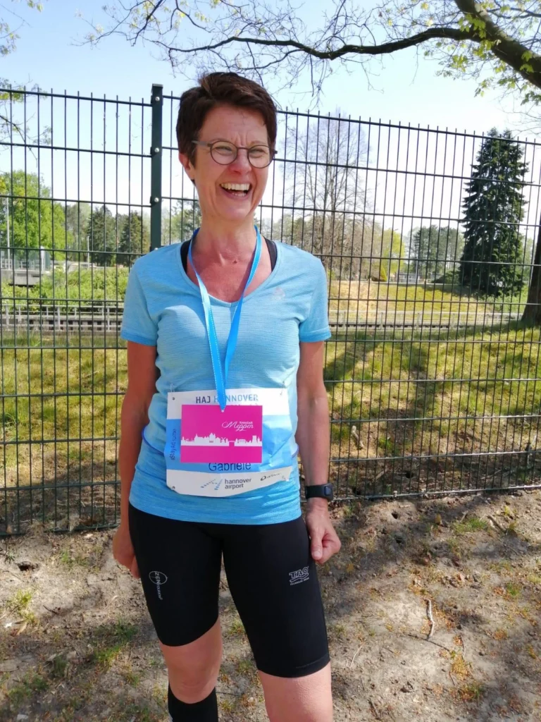 Lächelnde Frau in Sportkleidung mit Startnummer beim Hannover Halbmarathon, im Hintergrund ein Zaun und grüne Landschaft.