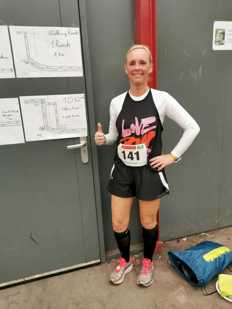 Teilnehmerin eines Laufwettbewerbs steht lächelnd vor einem Schild mit Streckeninformationen. Sie trägt ein sportliches Outfit mit der Startnummer 141 und hebt Daumen nach oben. Im Hintergrund sind Karten für den Walking- und Laufkurs sichtbar.