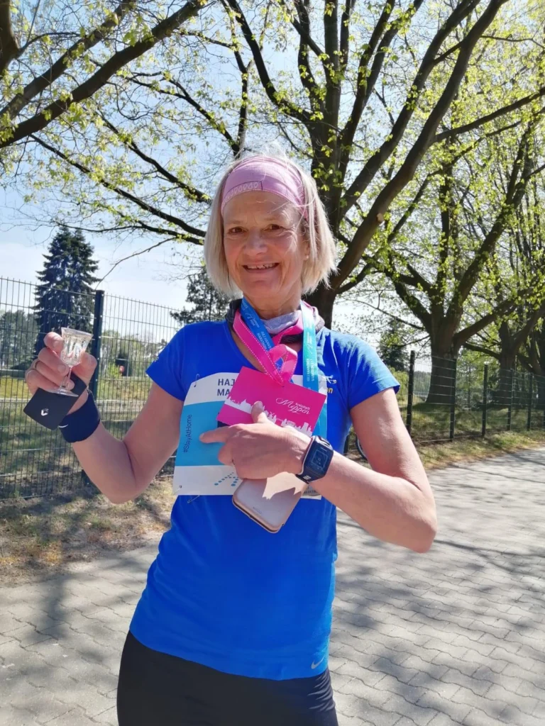 Läuferin mit Medaille und Urkunden in blauer Sportkleidung, steht im Freien unter Bäumen, lächelnd nach einem Wettkampf.