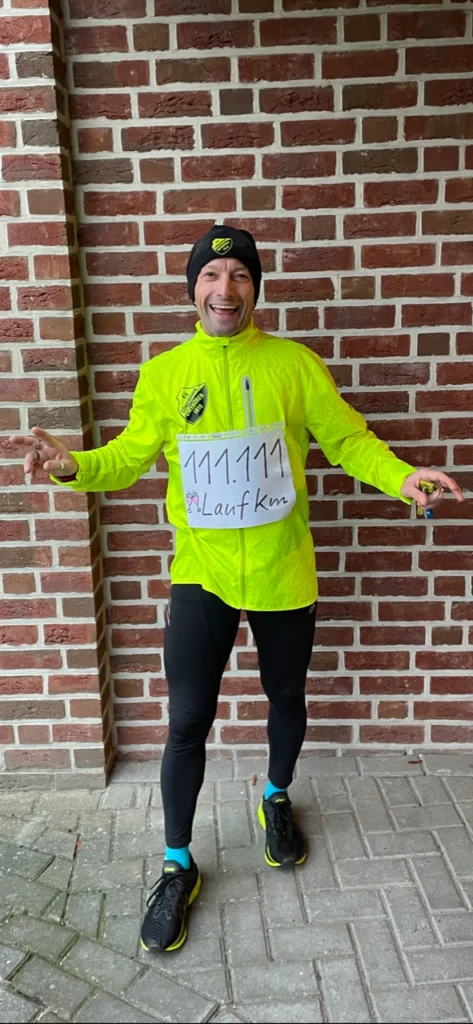 Läufer in leuchtend gelber Jacke und schwarzem Beanie, der vor einer Backsteinmauer steht. Er zeigt stolz die Nummer 111.111 auf seinem Laufshirt und posiert mit einer Handgeste.